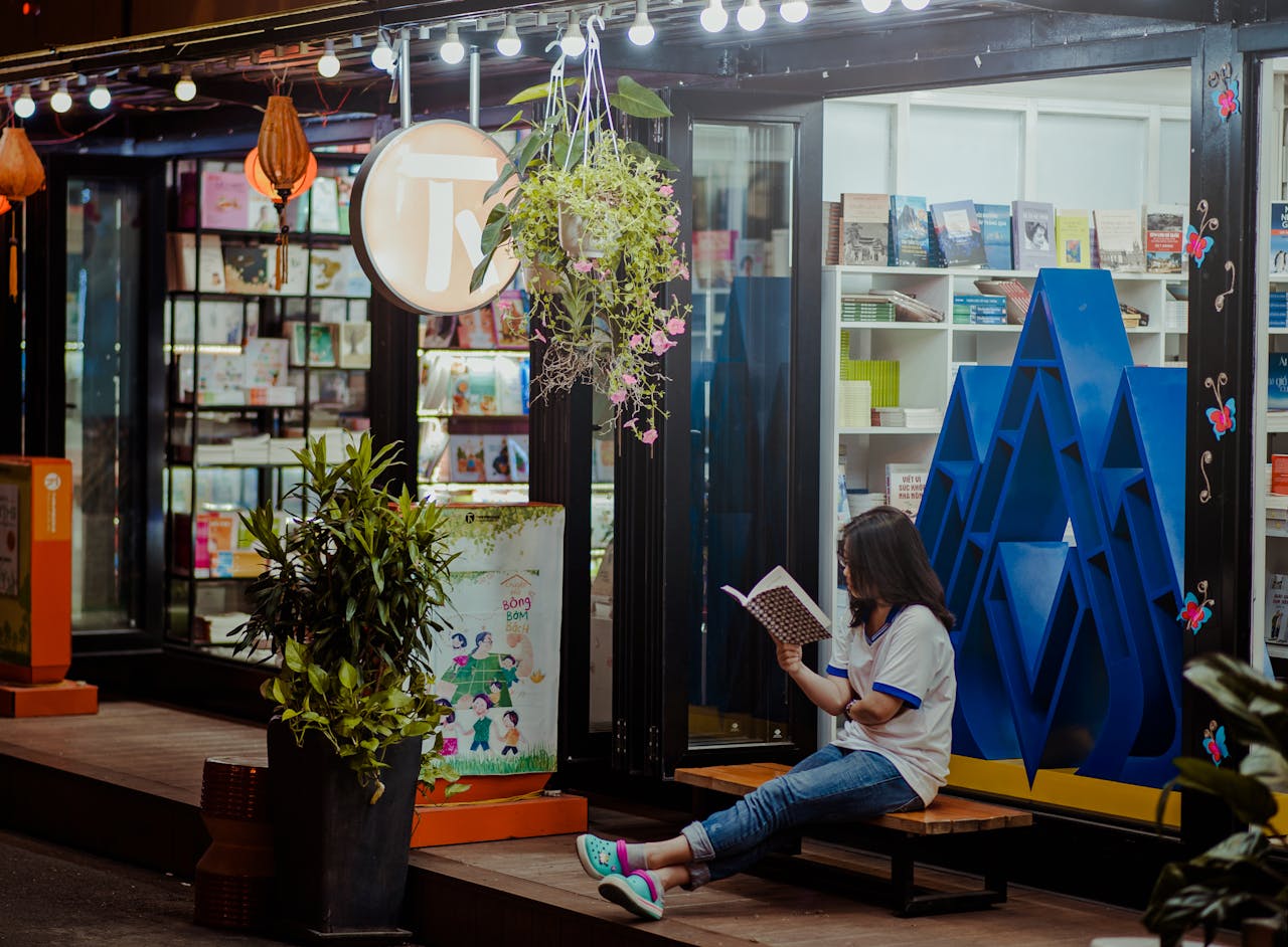 A woman sitting and reading outside a well-lit bookstore during the evening.